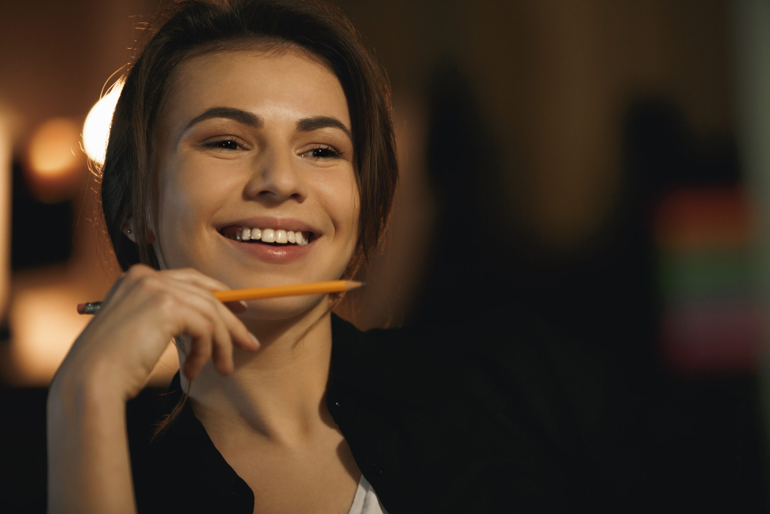 Photo of smiling young lady designer sitting indoors at night holding pencil. Looking aside.